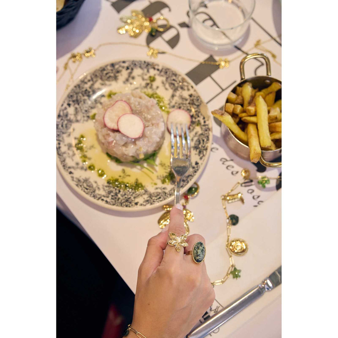 Main féminine portant une bague fleur dorée à 5 pétales avec perle blanche, accompagnée de bijoux en acier inoxydable sur une table de déjeuner chic.