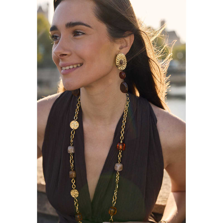 Femme portant des boucles d’oreilles dorées soleil en acier inoxydable avec perle orangée et pierre marron – Bijoux élégants et résistants à l’eau