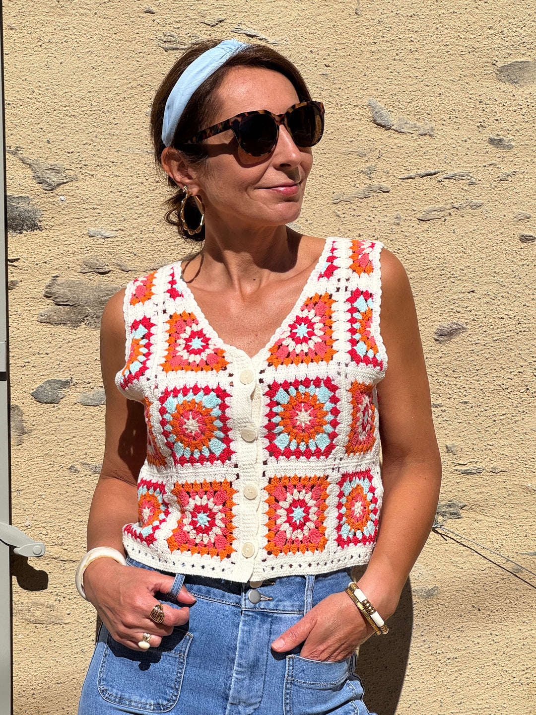 Gilet en crochet style bohème, sans manches avec col V, motifs colorés orange, rose, rouge et bleu ciel sur fond beige, parfait pour une tenue estivale tendance et légère.