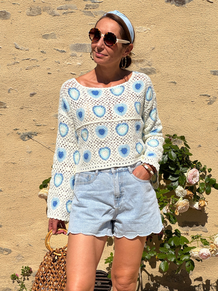 Top en crochet ajouré bleu et bleu ciel sur fond beige avec col bateau, manches longues et coupe courte, un haut léger au style bohème chic idéal pour l’été.