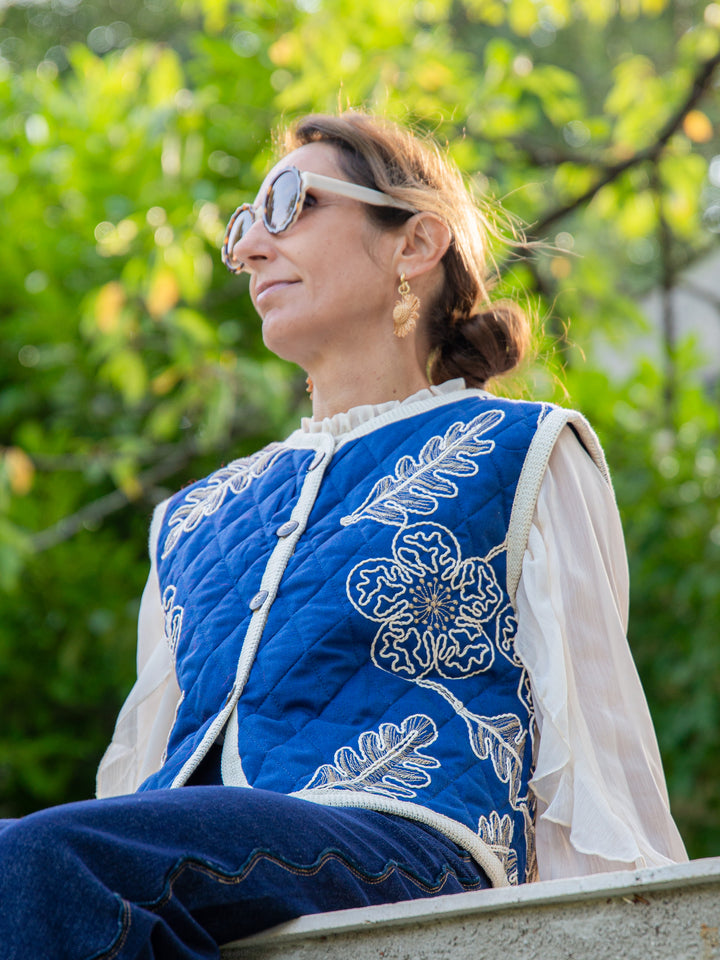 Femme portant une veste sans manche matelassée bleu brodée assise au soleil avec chemise écru